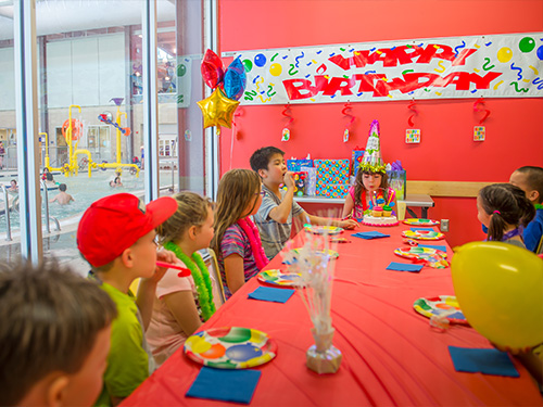 A group of children sitting around a long table covered in birthday decorations. One child is blowing candles out on a stack of cupcakes.