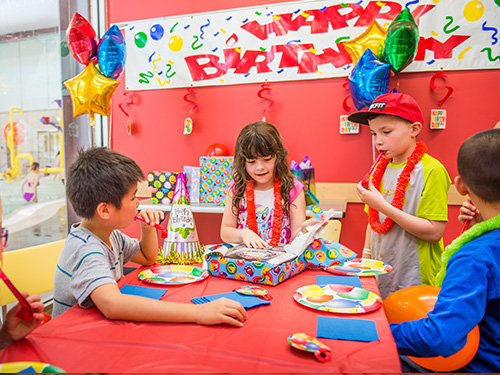 A child in a red party room opens a gift while three friends watch with excitement.