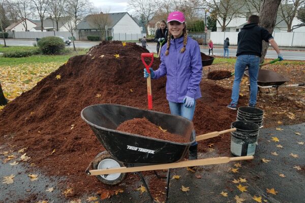 A volunteer in a purple jacket poses with a shovel next to a mulch pile and wheelbarrow