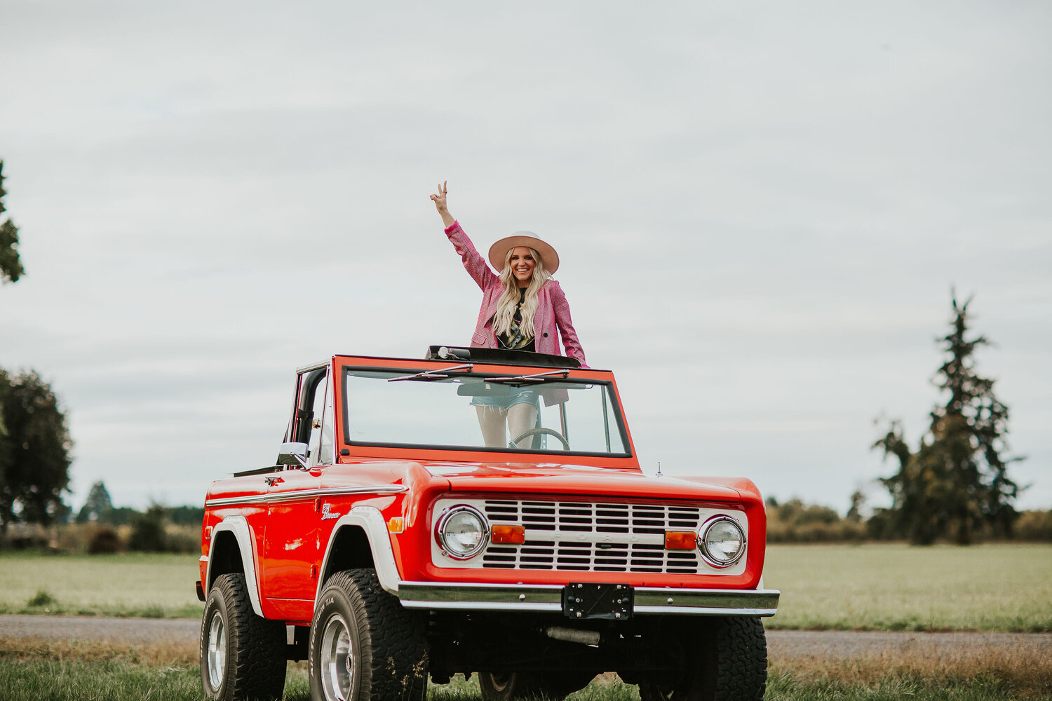 Musician, Britnee Kellogg poses with a outstretched arm, making a peace sign. She is standing in a red Jeep and is wearing pink leather jacket and wide brim hat.