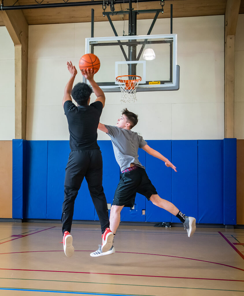 Two teens are playing basketball in an indoor gym. One is shooting the ball while the other is attempting to block. 