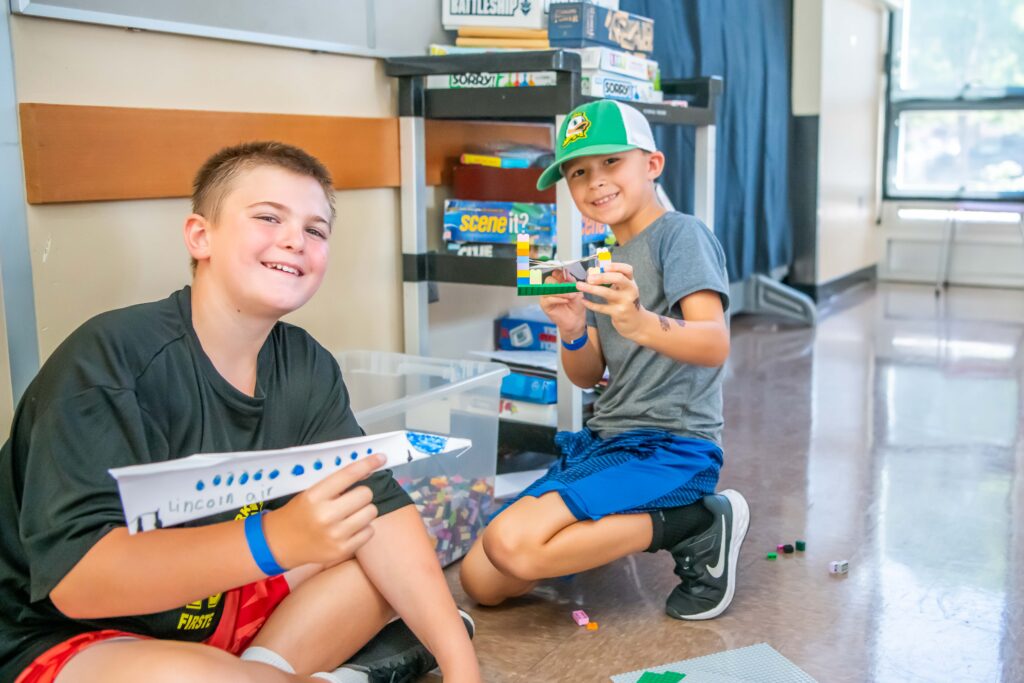 Two kids smile at the camera and show off their paper airplanes.