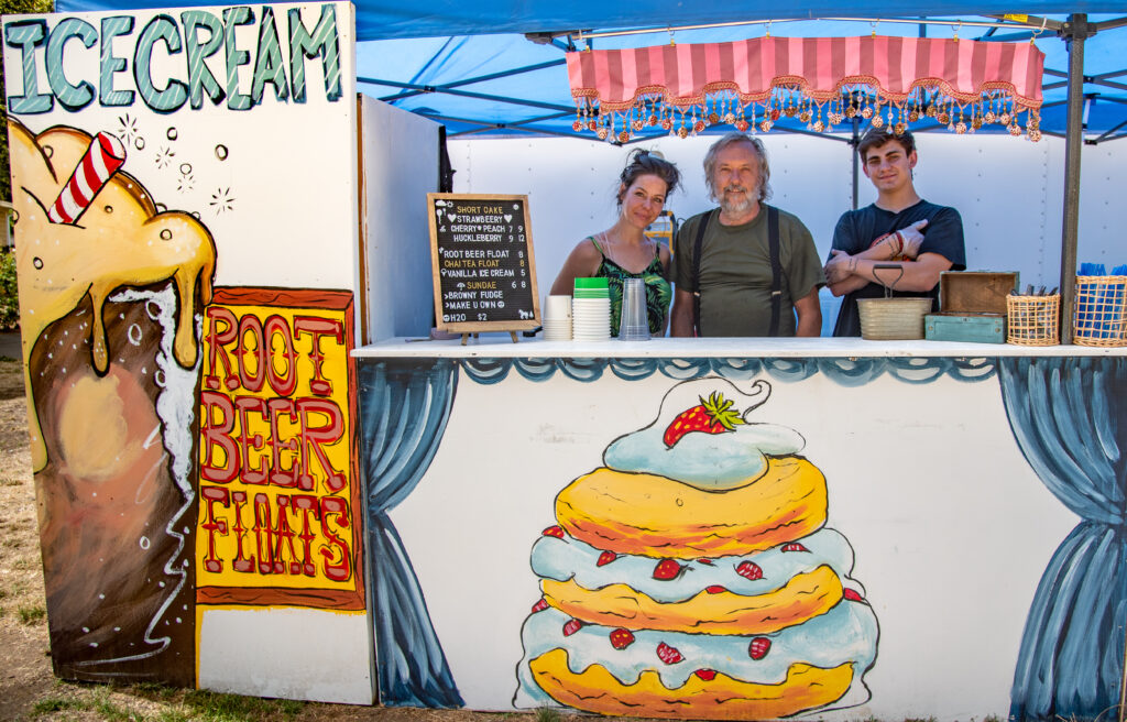 Three people working at an outdoor ice cream booth smile at the camera