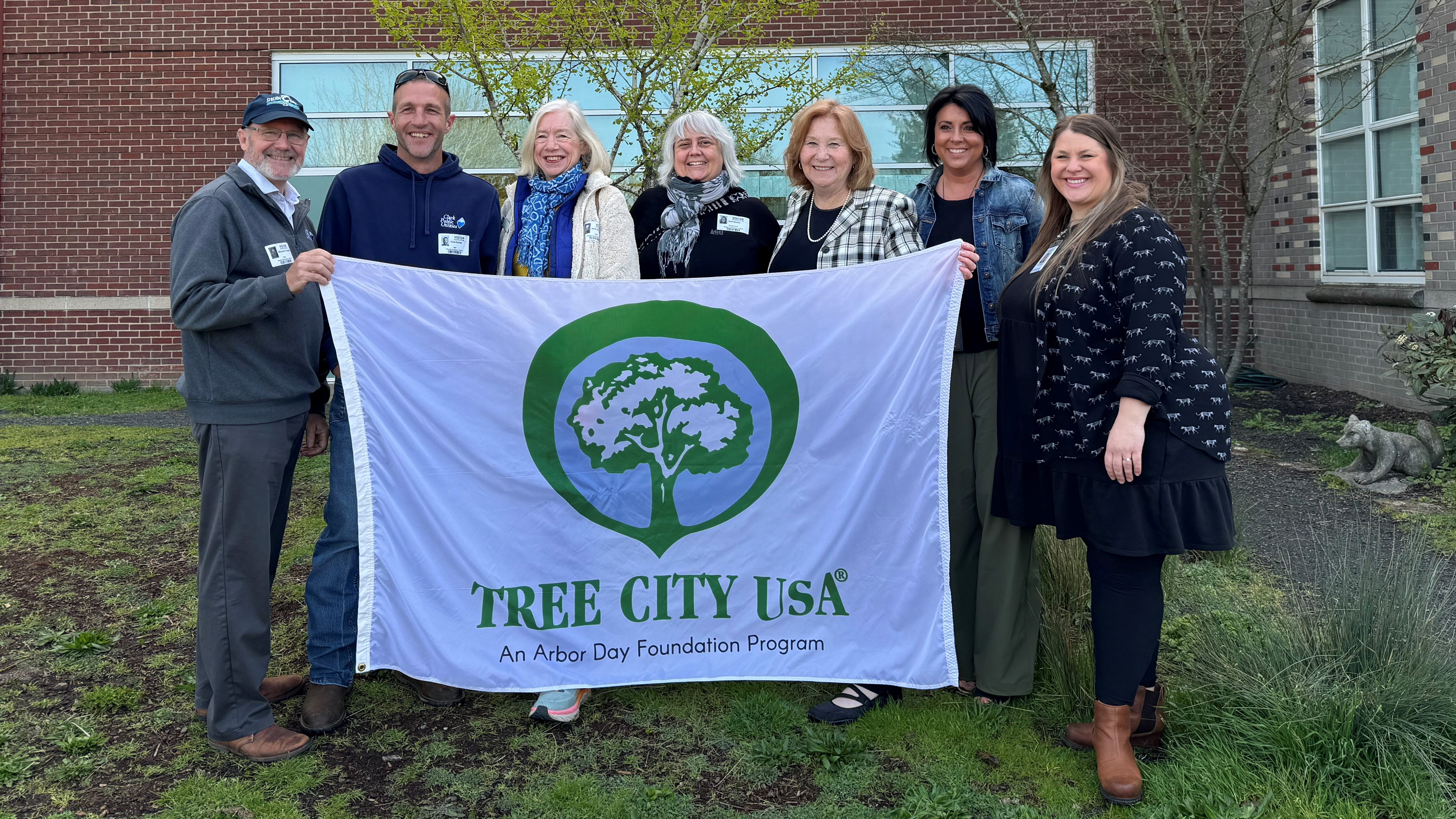 Seven people hold the Tree City USA Flag in front of Hough Elementary School. From left to right Clark Public Utilities partners, 2024 Mac Award Winner Jane Tesner Kleiner, Mayor Anne, teachers from Hough Elementary.